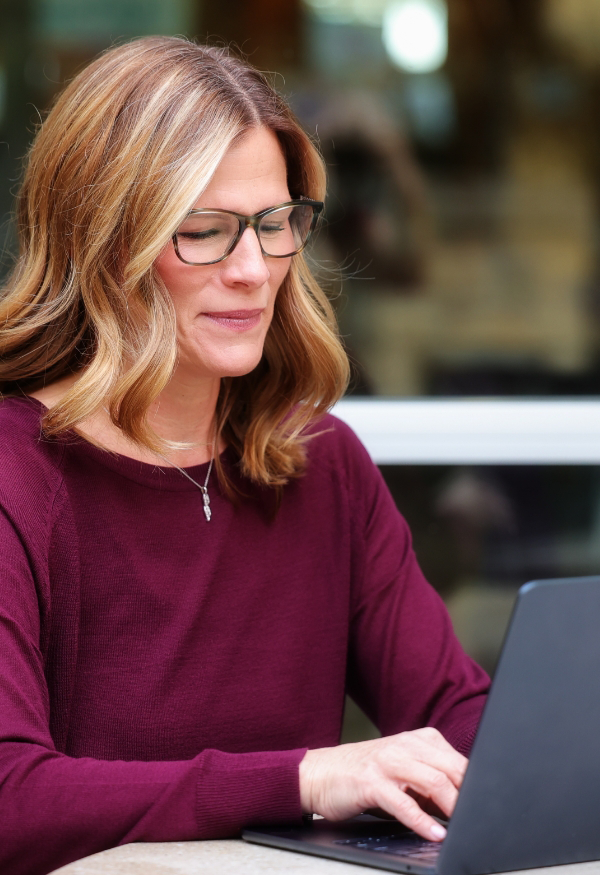 Ashley Ward with glasses and a burgundy sweater types on a laptop outdoors.