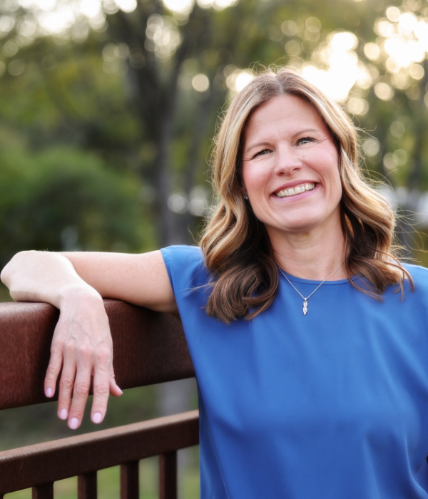 Ashley Ward in a blue shirt smiles, leaning on a wooden railing with trees in the background.