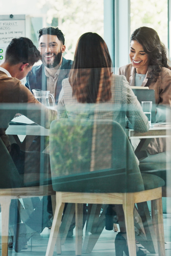 Four people in business attire sit around a table in a meeting room, engaged in discussion, with a flip chart in the background.