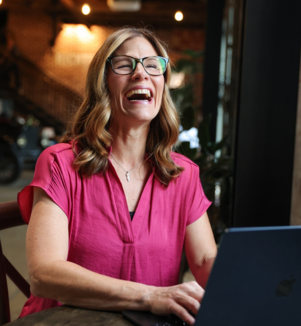 Ashley Ward in a pink shirt and glasses is laughing while working on a laptop in a cozy indoor setting.