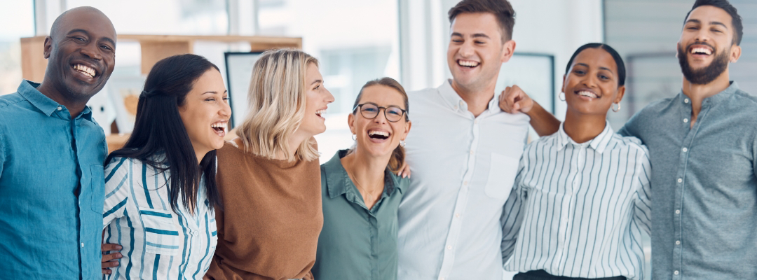 Group of seven diverse people standing close together indoors, all smiling and laughing.